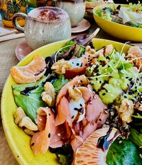 A beautiful cup of cappuccino with milk foam and chocolate sprinkled on it; healthy salad with salmon and orange slices; orange juice in beautiful jars; bright plate; nuts; healthy food 