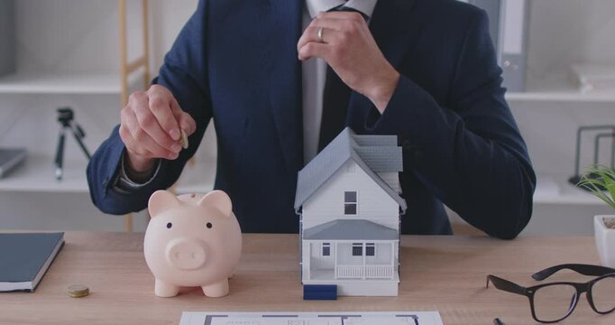 Cropped shot of businessman in suit saving money and putting coin inside piggy bank standing on desk next to small house. The concept of economy, budget and saving up money in order to buy own house.