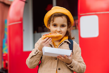 Cute little girl eating hot dog nearby food track on a street. Food delivery or take away food...