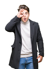 Young elegant man wearing winter coat over isolated background doing ok gesture shocked with surprised face, eye looking through fingers. Unbelieving expression.