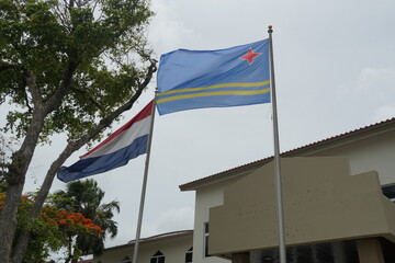 Aruba flag and Netherland flag are on metal poles and blow in the wind side by side in Oranjestad. There are some buildings and trees and overcast sky. There is a lot of copy space.