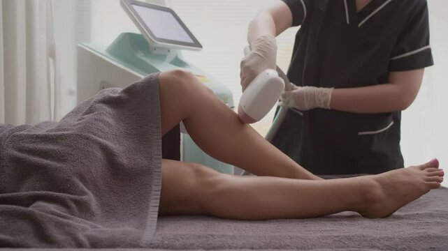 Tracking shot of unrecognizable beautician using IPV epilator on legs of client during procedure of hair removal at studio