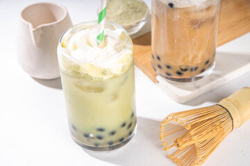 Classic and green matcha boba bubble tea with tapioca pearls and whipped non-dairy creamer on top of glass, summer cold sweet dessert drink on white table background copy space