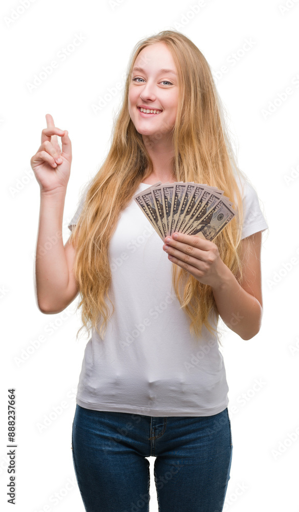 Sticker blonde teenager woman holding dollars surprised with an idea or question pointing finger with happy 