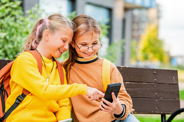two charming schoolgirls look at the smartphone screen while sitting on bench on a city street.