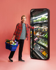 Man in casual clothes, holding grocery basket, reaching into huge smartphone screen showing store...