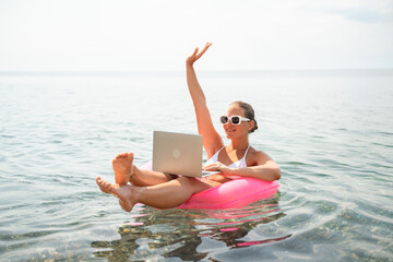 A woman is sitting on a pink inflatable raft in the ocean, holding a laptop. She is smiling and she is enjoying her time. Concept of relaxation and leisure.