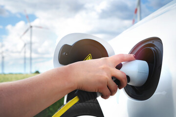 woman's hand with painted nails plugging a charger cable into an electric vehicle. Close-up with wind power plants blurred in background. sustainable transportation concept. green energy, eco-friendly