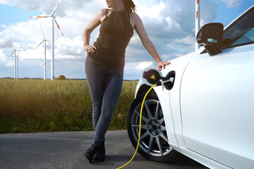 EV charging in sunny scenery with wind turbines in background. White car. Yellow cable. Beautiful...