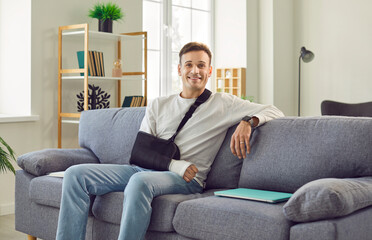 Happy man wearing orthopedic sling on injured arm sitting on comfortable sofa at home, looking at...