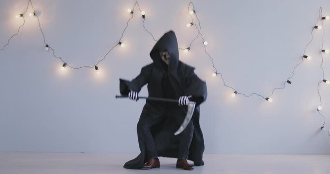 An actor in a black Grim Reaper costume dances funny against a background of light bulbs. Halloween party concept, fun dancer in death suit. Costume accessories 