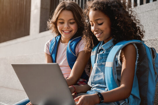 Two happy elementary middle school girls friends pupils classmates using laptop gadgets looking on screen doing homework project together sitting on the stairs steps outdoors