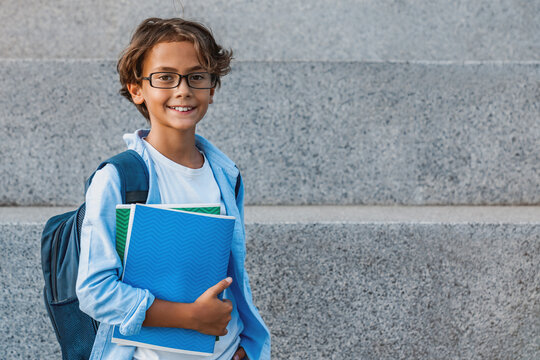 Portrait of happy caucasian young preteen boy elementary middle school pupil in eyeglasses with backpack holding notebook books outside the primary school. Education concept copy space