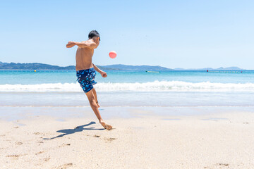 A young boy in blue shorts energetically kicks a red ball on a sunny beach, with clear blue waters and distant mountains in the background. 