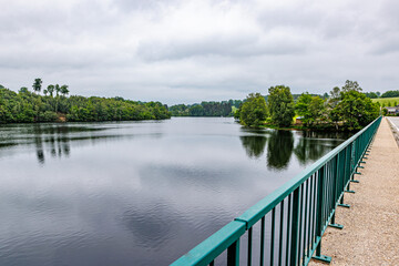 Fototapeta premium Lake Robertville with its calm waters seen from vehicular bridge with pedestrian sidewalk, slight reflection on water surface, green leafed pine trees in background, cloudy day in Waimes, Belgium