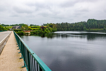 Fototapeta premium Lake Robertville and vehicular bridge with pedestrian sidewalk, slight reflection on water surface, hills with green trees in background, cloudy day in Waimes, Belgium
