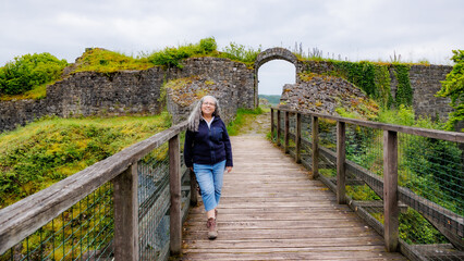 Smiling senior adult woman tourist standing on a wooden bridge leading to an arched door at Logne Castle, time-worn stone wall against gray cloud-covered sky, cloudy day in Ferrieres, Belgium