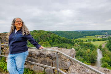 Naklejka premium Smiling female tourist leaning on metal railing at ruins of Logne castle on hill, Ourthe river, farmland and lush green trees in background, blue casual clothing, cloudy day in Ferrieres, Belgium