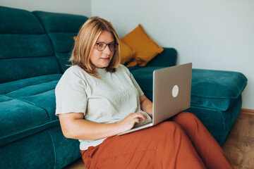 Nice beautiful woman in glasses working at the notebook near the sofa at home. Concepts of...