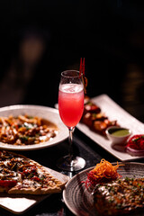 Red cocktail drink in glass on dark background with foods on table. Pizza, Pasta and Kebab platter with Cocktail.