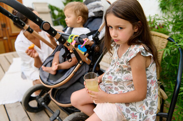 Obraz premium Young girl holding a glass of juice while sitting next to a baby in a stroller and mother. Scene captures outdoor family time and sibling interaction.