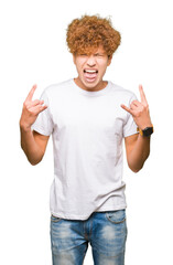 Young handsome man with afro hair wearing casual white t-shirt shouting with crazy expression doing rock symbol with hands up. Music star. Heavy concept.