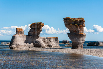 Breathtaking limestone sculptures rise from a shallow coastal pool at Mingan Archipelago National Park Reserve, framed by a clear blue sky adorned with dramatic clouds.