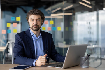 Fototapeta premium Confident businessman working on laptop in modern office with glass walls. Professional setting for corporate planning, strategy, or execution.