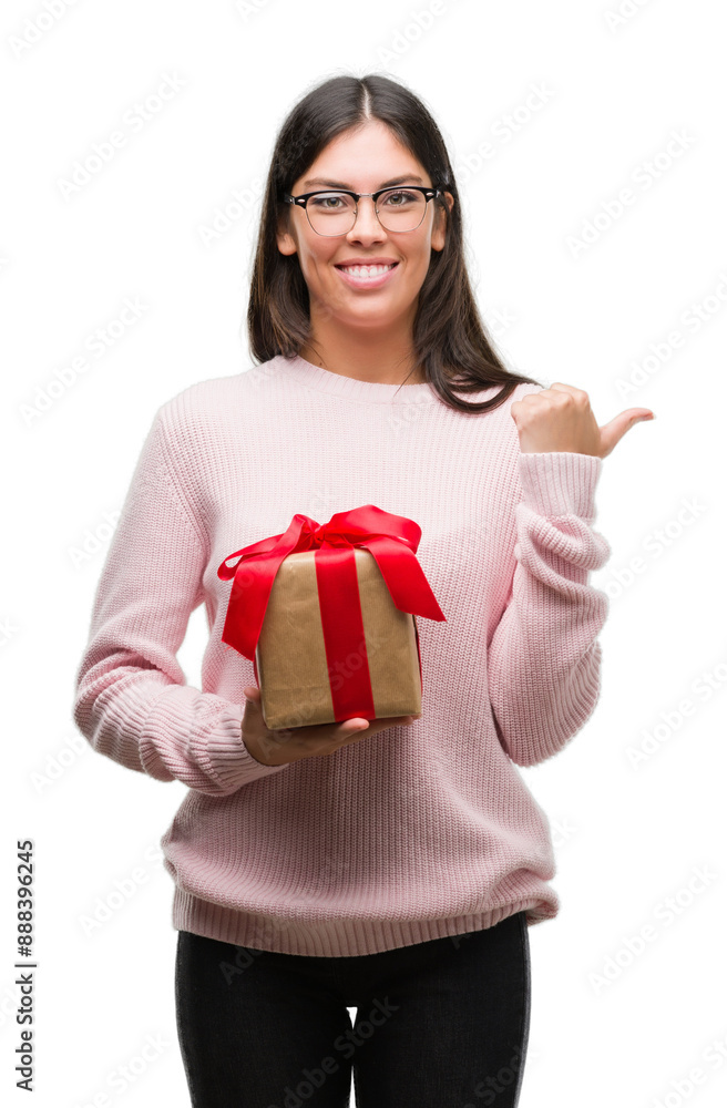 Wall mural young hispanic woman holding a gift pointing and showing with thumb up to the side with happy face s