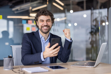 Cheerful businessman celebrating success in modern office. Man in suit holding smartphone, smiling,...