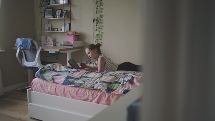 Young girl rests and lies on cozy bed in big comfort room. Caucasian teen uses digital tablet computer and stylus to watch videos and surf Internet. Attractive teenager spends time in bedroom at home.
