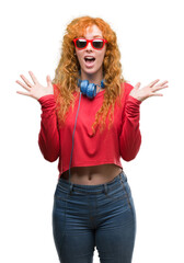 Young redhead woman wearing headphones very happy and excited, winner expression celebrating victory screaming with big smile and raised hands