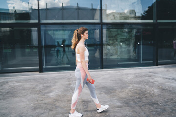 Young Caucasian woman dressed in athletic wear, walking outdoors while holding smartphone and listening to music with earphones. appears deep in thought, reflecting or planning in physical activity