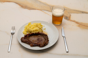 a grilled beef entrecote on a white plate with a little mashed potato, on a white veined marble table, next to a knife, fork and a glass with cold beer