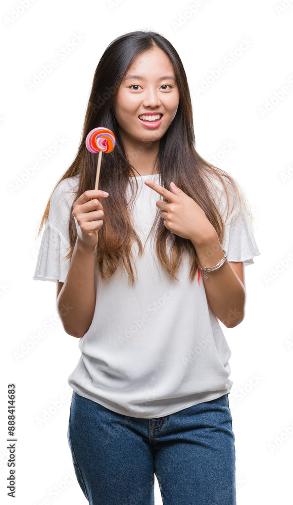 Poster Young asian woman eating lollipop candy over isolated background very happy pointing with hand and finger