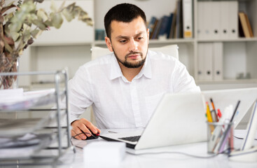 Fototapeta premium Male bookkeeper sitting at table and using laptop while doing his job in office.