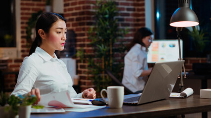 Asian bookkeeper working in office overnight, bored of checking financial data graphs and charts. Businesswoman trying to finish tedious paperwork, feeling stressed and extenuated