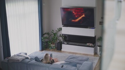 Adult woman sits on couch in cozy modern living room near TV and surfing internet using mobile phone. Caucasian woman rests, spends weekend at home. Flat screen TV monitor with displayed action movie.