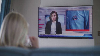 Adult woman sits on couch in living room and watches TV news about elections in United States of...