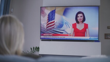 Adult woman rests on couch in cozy living room and watches TV news about national election day in...