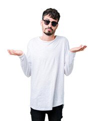 Young handsome man wearing sunglasses over isolated background clueless and confused expression with arms and hands raised. Doubt concept.