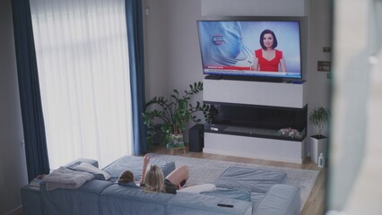 Adult woman rests on couch in modern living room and watches TV news about US elections. Young girl comes into room and sits on sofa. Big TV screen with displayed live news broadcast. High angle.
