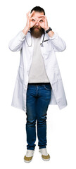 Young blond doctor man with beard wearing medical coat doing ok gesture like binoculars sticking tongue out, eyes looking through fingers. Crazy expression.