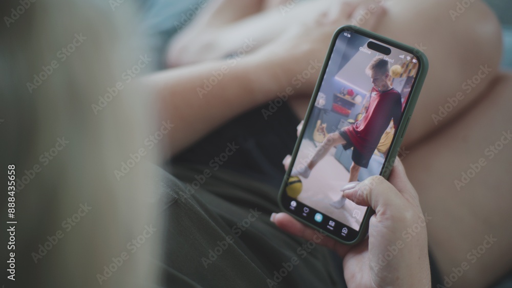 Poster caucasian woman scrolls through and watches app feed with short videos using mobile phone while lyin