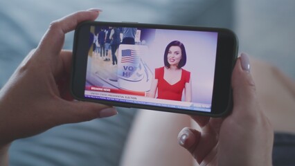Broadcast of TV news displayed on mobile phone. Close up of woman watching online breaking news about elections in the United States using smartphone while lying on sofa at home. Election Day in USA.