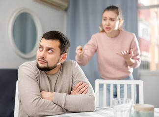 Fototapeta premium Sad man sitting at table in apartment while his wife standing behind and quarreling with him.