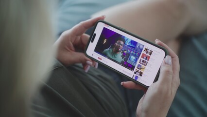Close up of Caucasian woman watching video about color grading using mobile phone while lying on sofa. Video hosting website with video content about color correction displayed on smartphone screen.