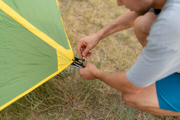 a person is kneeling on the ground, securing a tent peg to the ground near a tent. He is camping in...