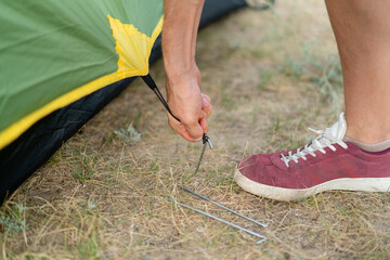 a person is kneeling on the ground, securing a tent peg to the ground near a tent. He is camping in a field with a body of water in the background © Mihail