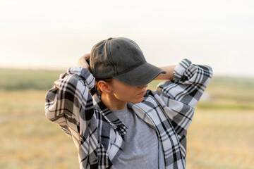 woman wearing a plaid shirt and stands in a field. She puts on a black cap and adjusting it  with one hand. Warm sunset sunlight outdoors in the field